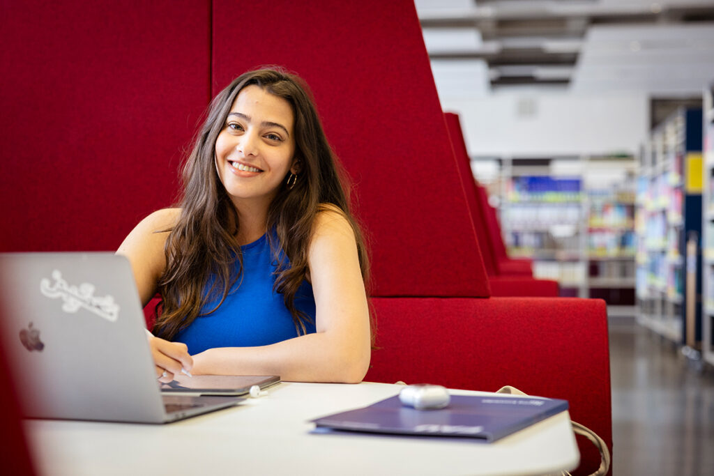 Eine Frau sitzt in der Erlanger Universitätsbibliotek auf einem gepolsterten Sitzmöbel vor ihrem Laptop und lächelt in die Kamera.